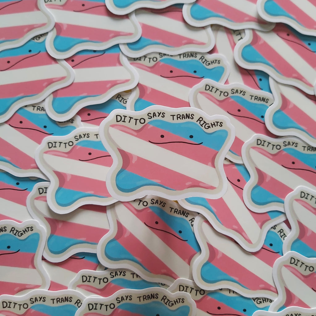 A pile of die-cut stickers shaped like a transgender pride flag, featuring pink and light blue stripes with a white border. One sticker in the foreground reads 'DITTO SAYS TRANS RIGHTS' in black text.