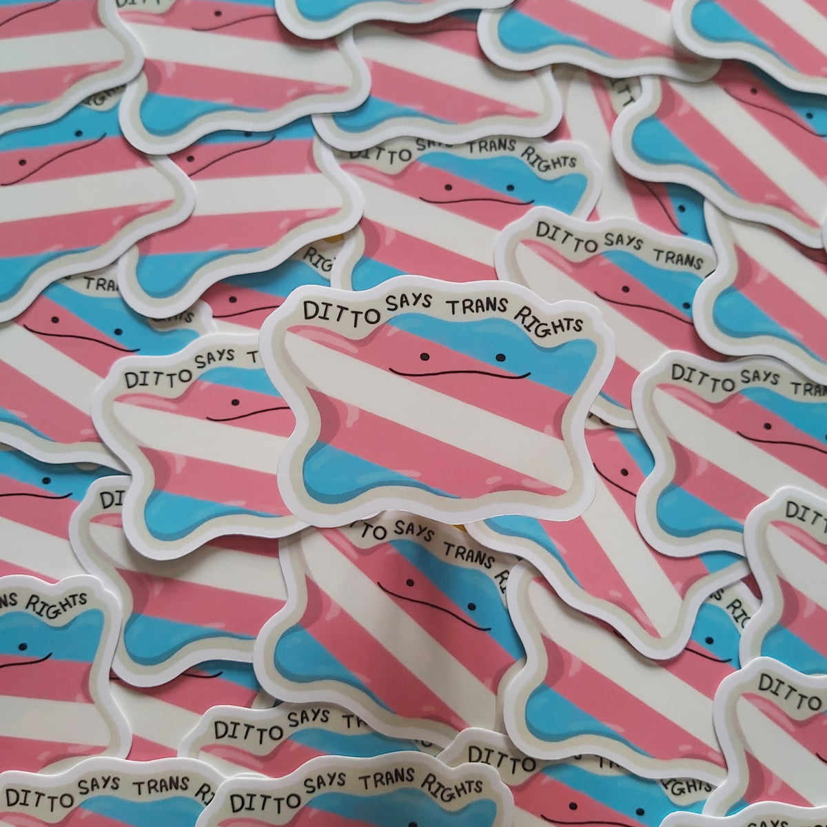 A pile of die-cut stickers shaped like a transgender pride flag, featuring pink and light blue stripes with a white border. One sticker in the foreground reads 'DITTO SAYS TRANS RIGHTS' in black text.