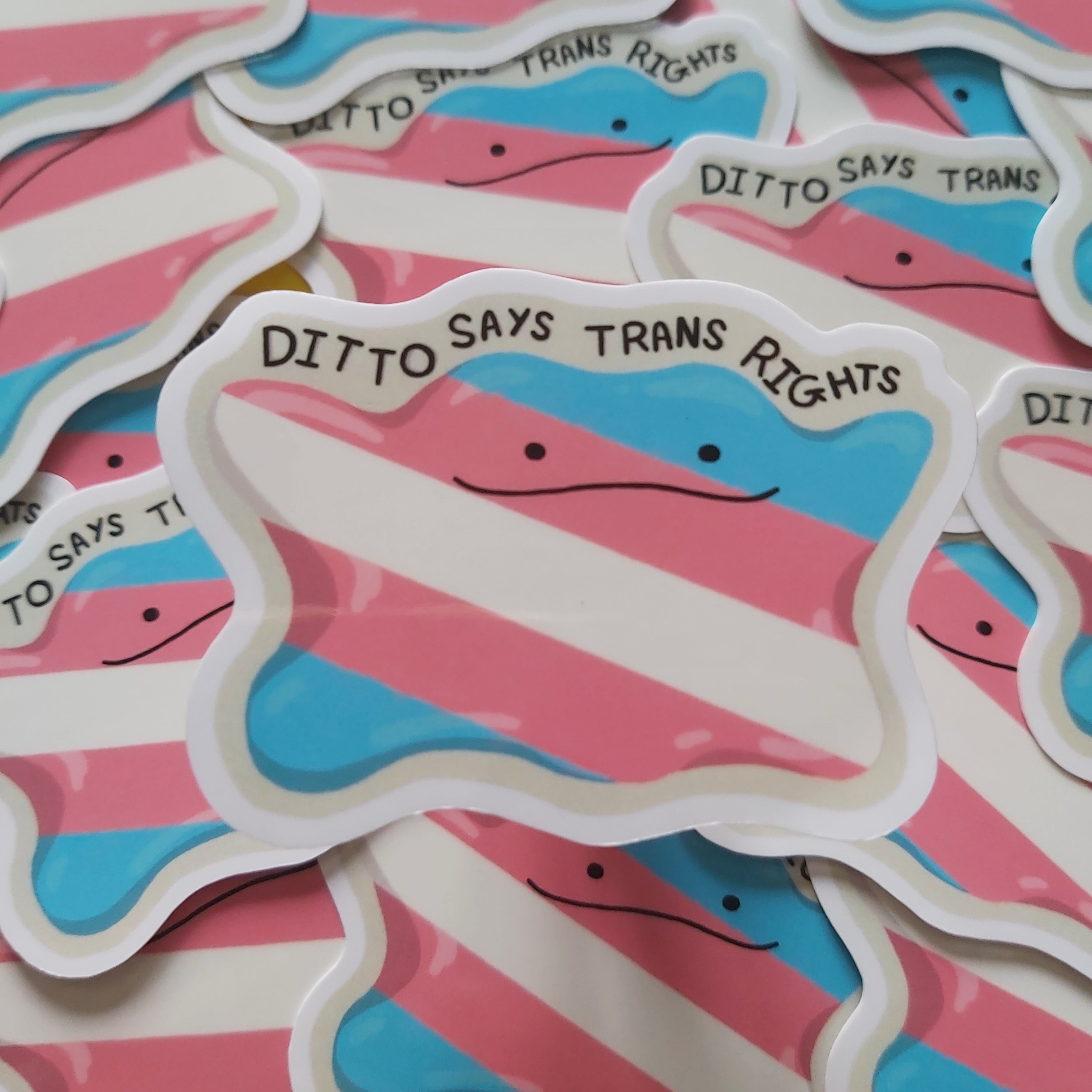 A pile of die-cut stickers shaped like the transgender pride flag, with the text 'DITTO SAYS TRANS RIGHTS' written across the top. The stickers feature the pink, white, and blue stripes of the transgender pride flag with a simple ditto face.