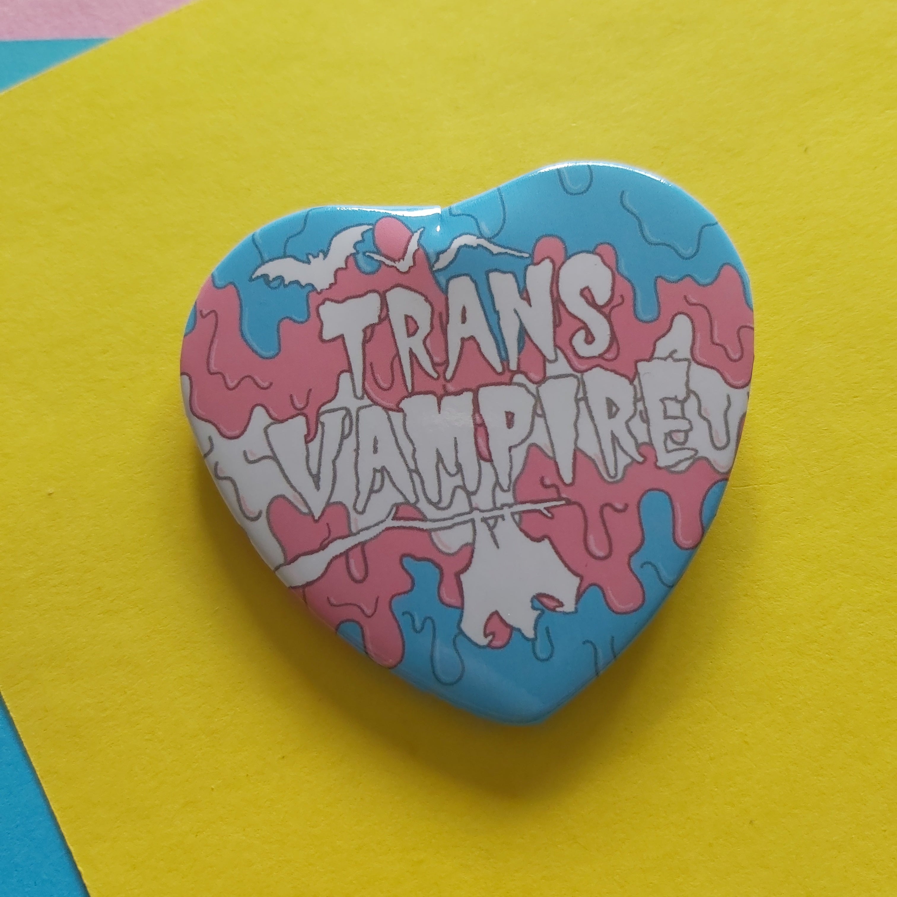 A heart-shaped pin featuring a pink and blue paint-drip design with the words 'TRANS VAMPIRE!' in white stylised lettering, displayed on a bright yellow paper background.