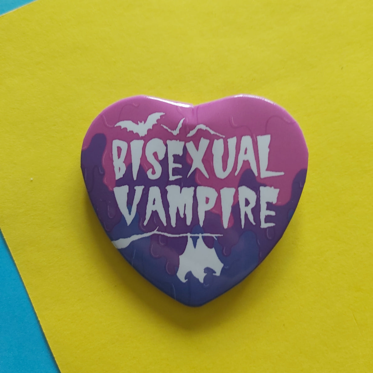 A heart-shaped pin with a pink and purple gradient background, featuring the text 'BISEXUAL VAMPIRE' in white stylised lettering, along with bat silhouettes and a white bat silhouette at the bottom, displayed on a bright yellow paper background.