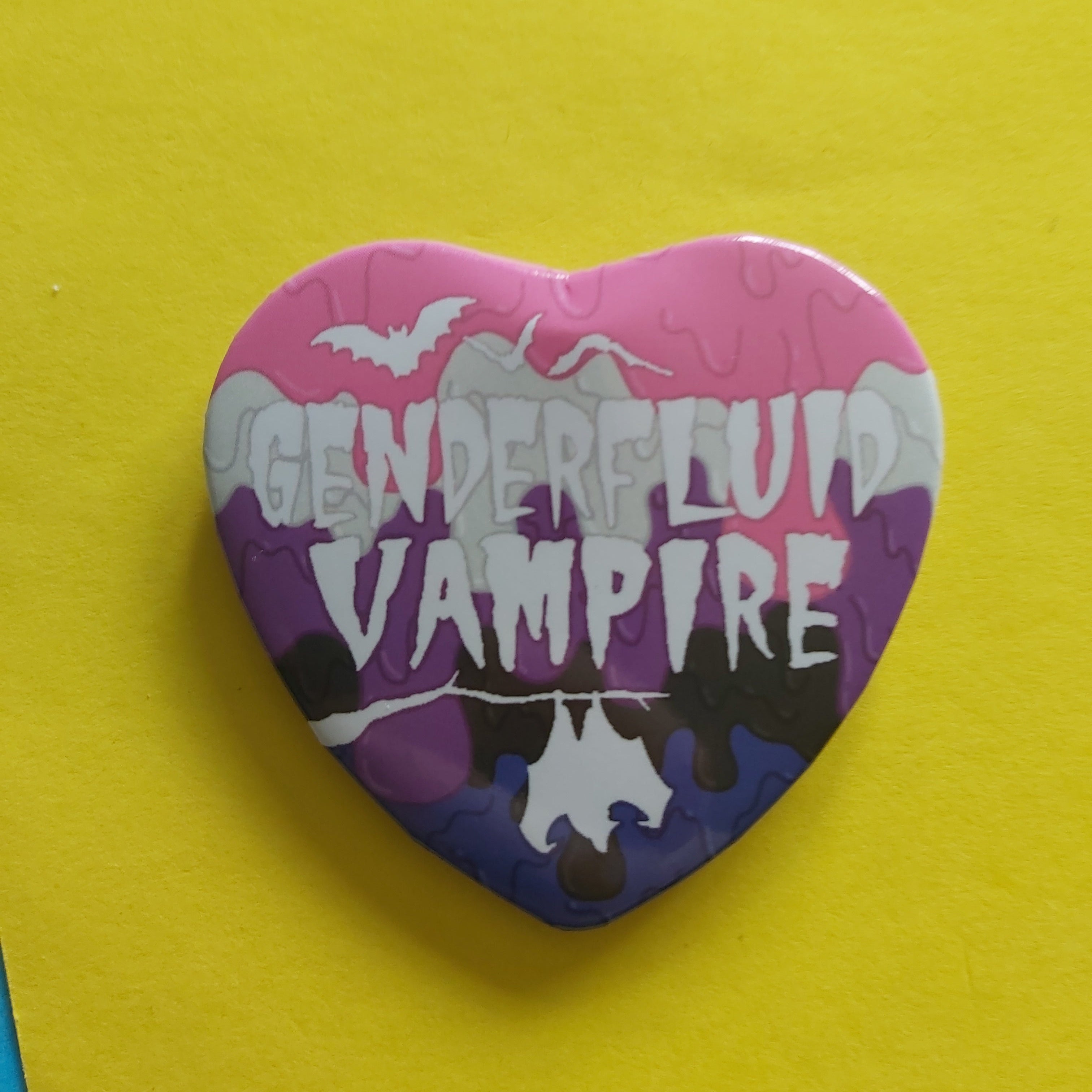 A heart-shaped pinback button featuring a multicolour paint-dripping design with the text 'GENDERFLUID VAMPIRE' in white, set against a bright yellow background.