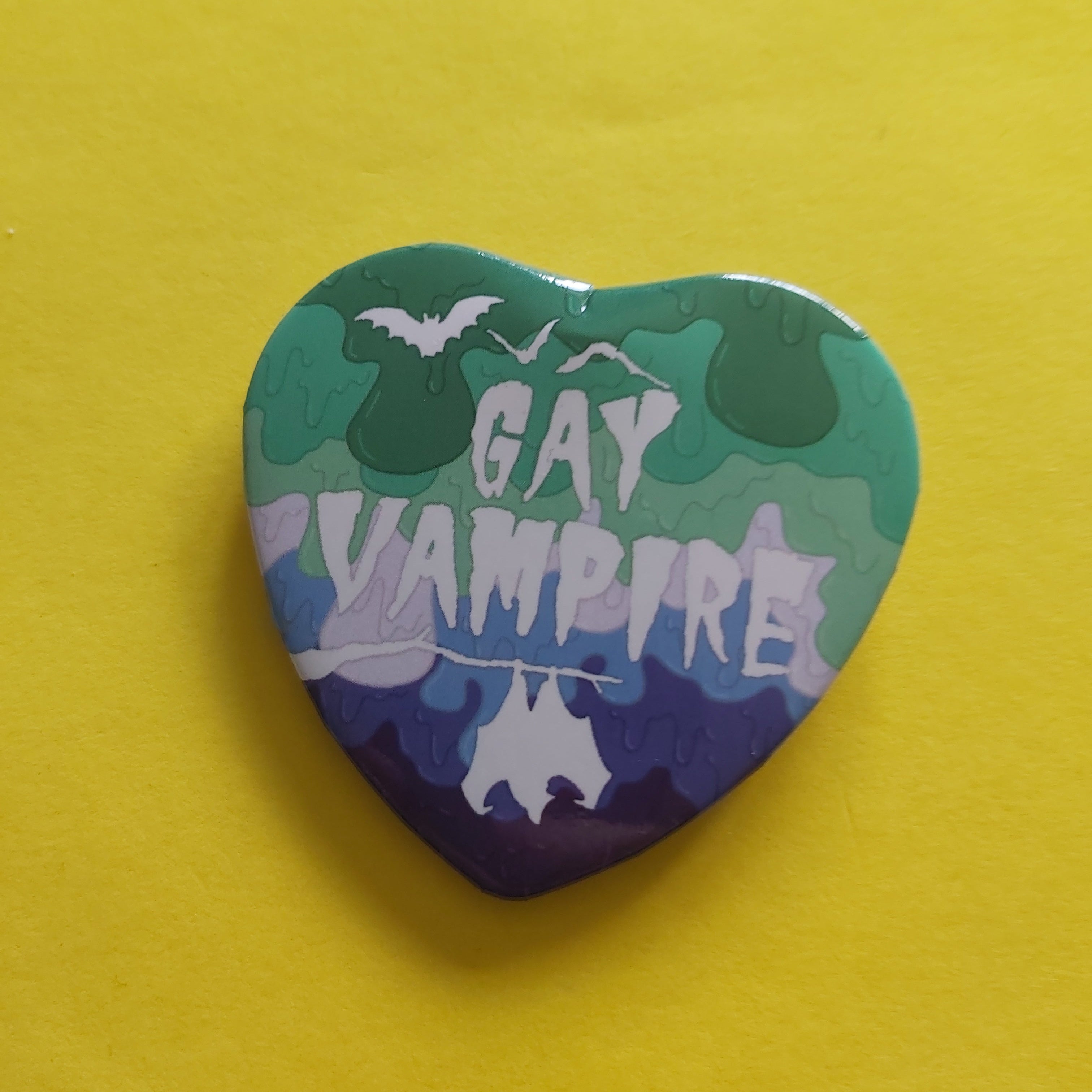 A heart-shaped pin featuring a green, blue, and white gradient with bats and the words 'GAY VAMPIRE' in white, displayed on a bright yellow background.