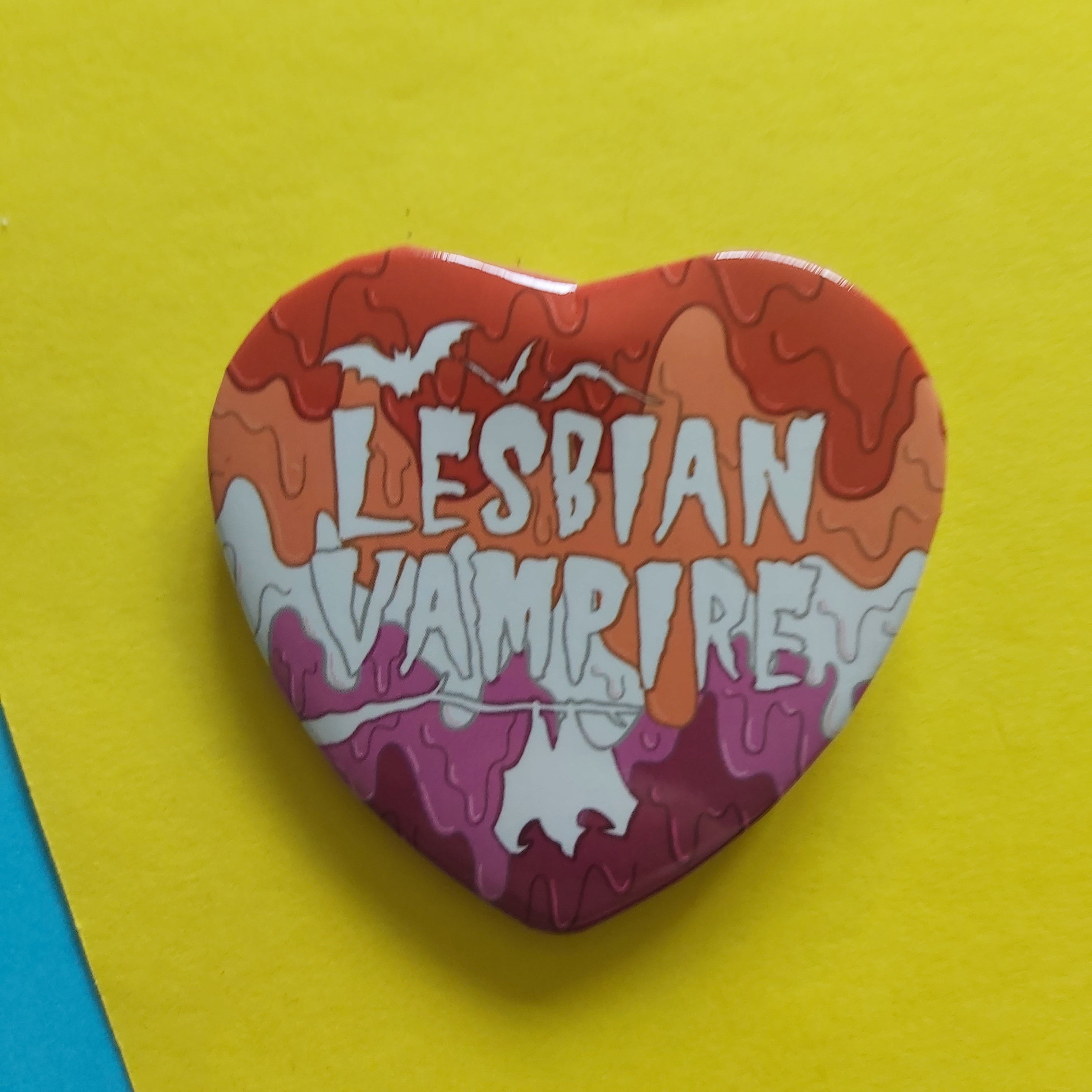 A heart-shaped pin with a dripping paint design in orange, peach, and pink, featuring the text 'LESBIAN VAMPIRE' in white. The pin is photographed on a bright yellow background with a blue corner visible in the lower-left.