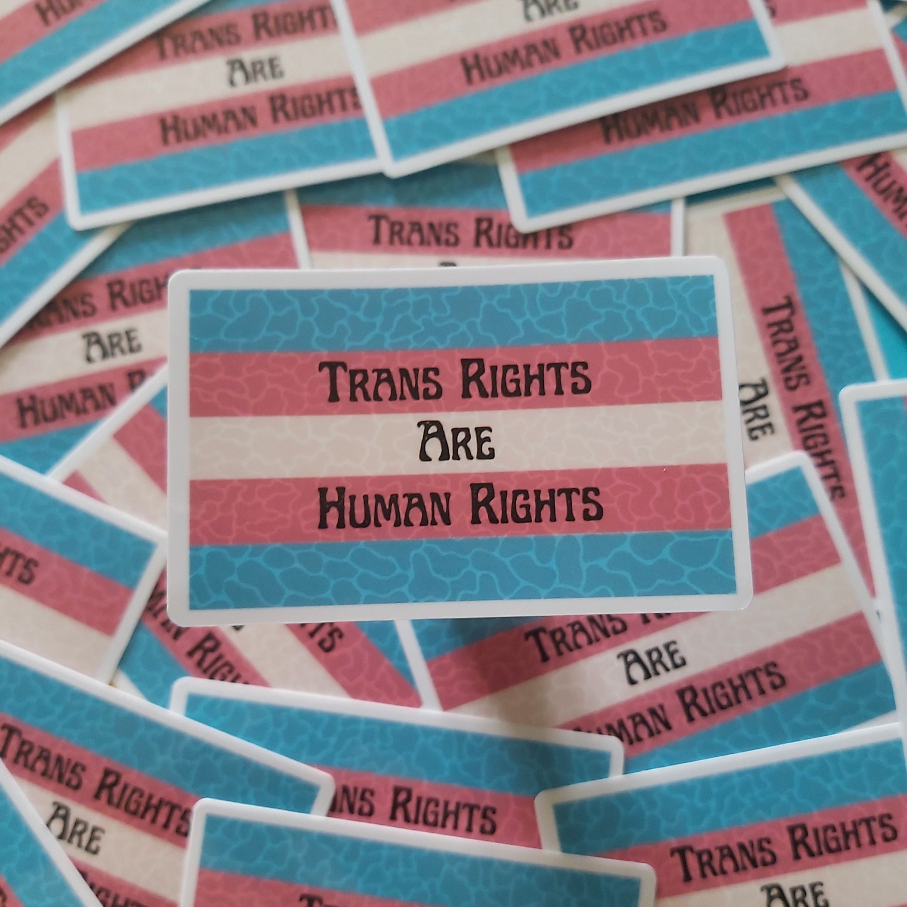 A pile of rectangular stickers featuring the text 'TRANS RIGHTS ARE HUMAN RIGHTS' in black lettering over horizontal red, white, and blue stripes, with a subtle blob pattern on the sections.