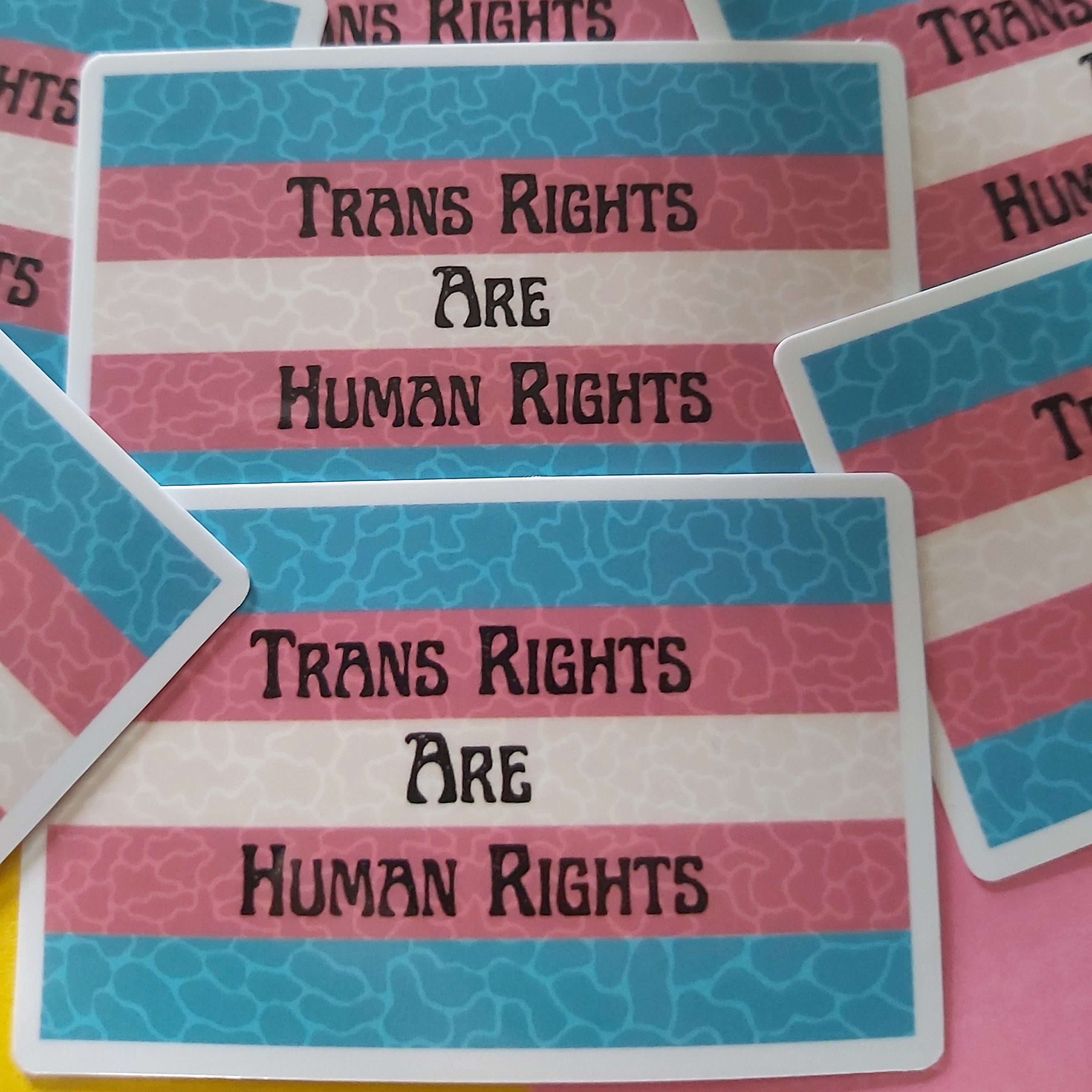 A close-up of several rectangular stickers stacked on top of each other, each printed with the phrase 'TRANS RIGHTS ARE HUMAN RIGHTS' in black text over horizontal blue, pink, and off-white stripes.