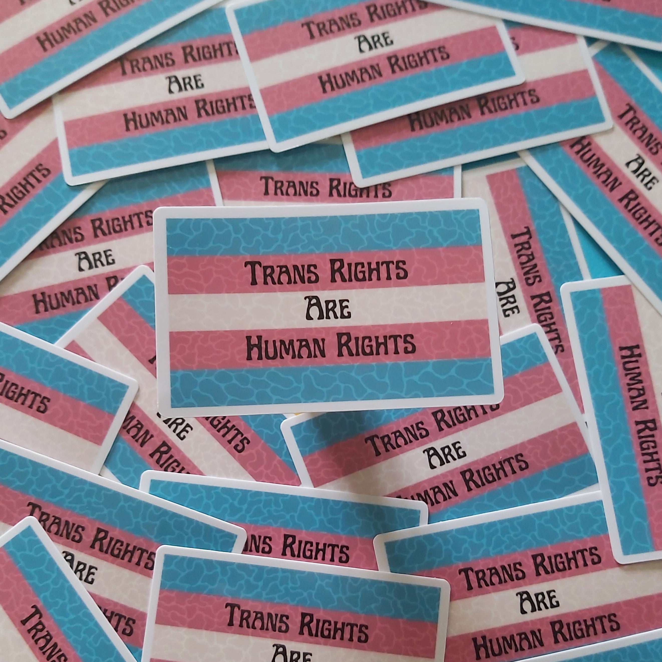 A pile of small rectangular stickers with the text 'Trans Rights Are Human Rights' printed in black on pink and blue striped backgrounds