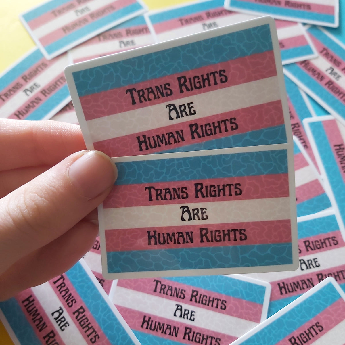 A hand holds two rectangular stickers with the text 'TRANS RIGHTS ARE HUMAN RIGHTS' in black lettering over horizontal pink, white, and blue stripes. Multiple identical stickers are scattered in the background on a bright pastel surface.