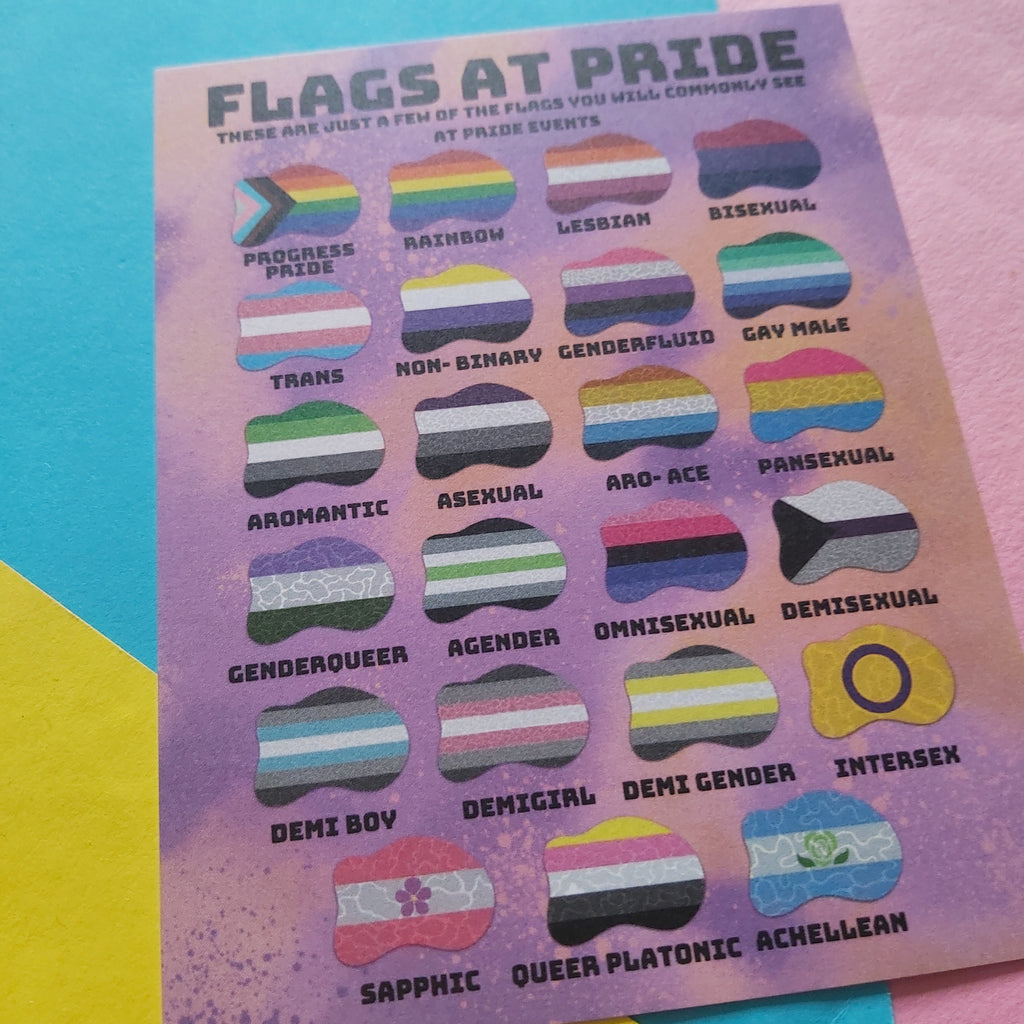 A printed card titled "FLAGS AT PRIDE" showing many small lesbian and transgender pride flag icons with labels, placed on colorful pastel paper background.