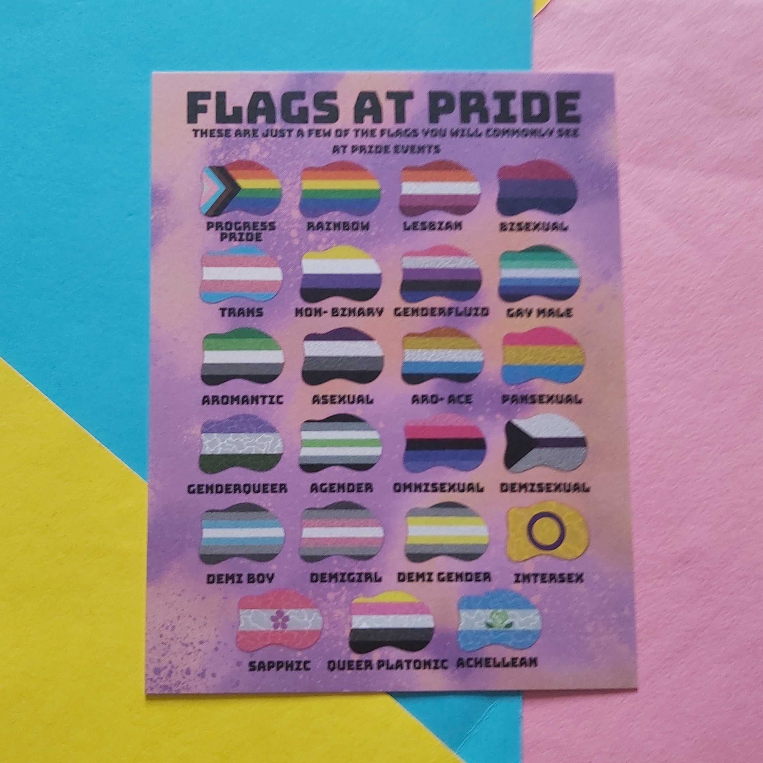 A printed card titled "FLAGS AT PRIDE" showing multiple small striped pride flags arranged in a grid on a pastel purple background, placed on top of bright colored paper shapes.