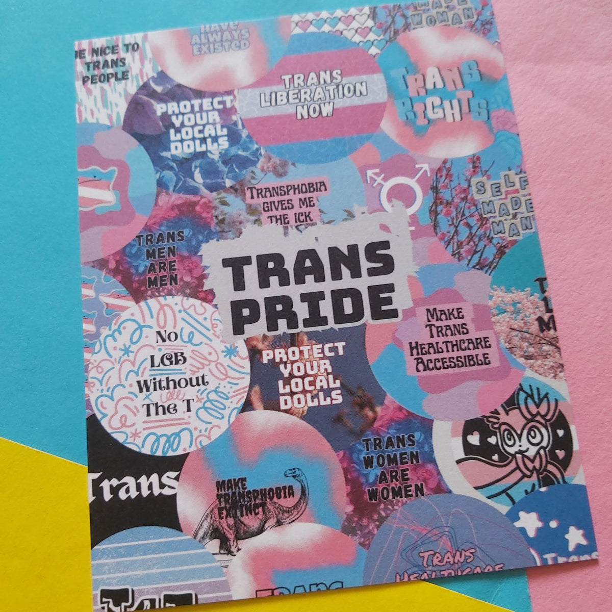 A print with a colourful collage design and the large text 'TRANS PRIDE' in the center, photographed on a bright pastel background with pink, blue, and yellow sections.