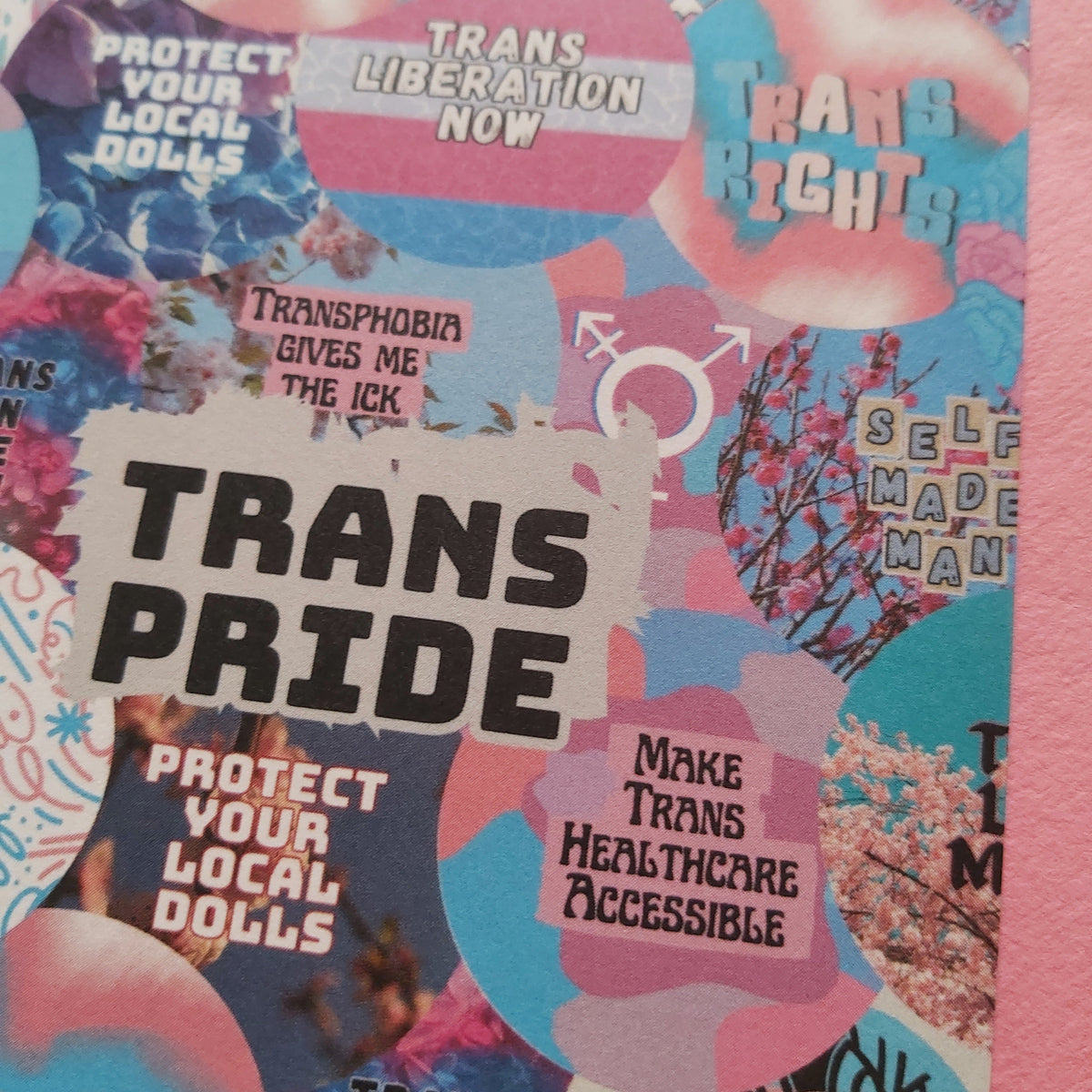 A close-up of a Trans Pride-themed collage print featuring pink, blue, and white shapes with text such as 'TRANS PRIDE,' 'PROTECT YOUR LOCAL DOLLS,' and 'MAKE TRANS HEALTHCARE ACCESSIBLE.'