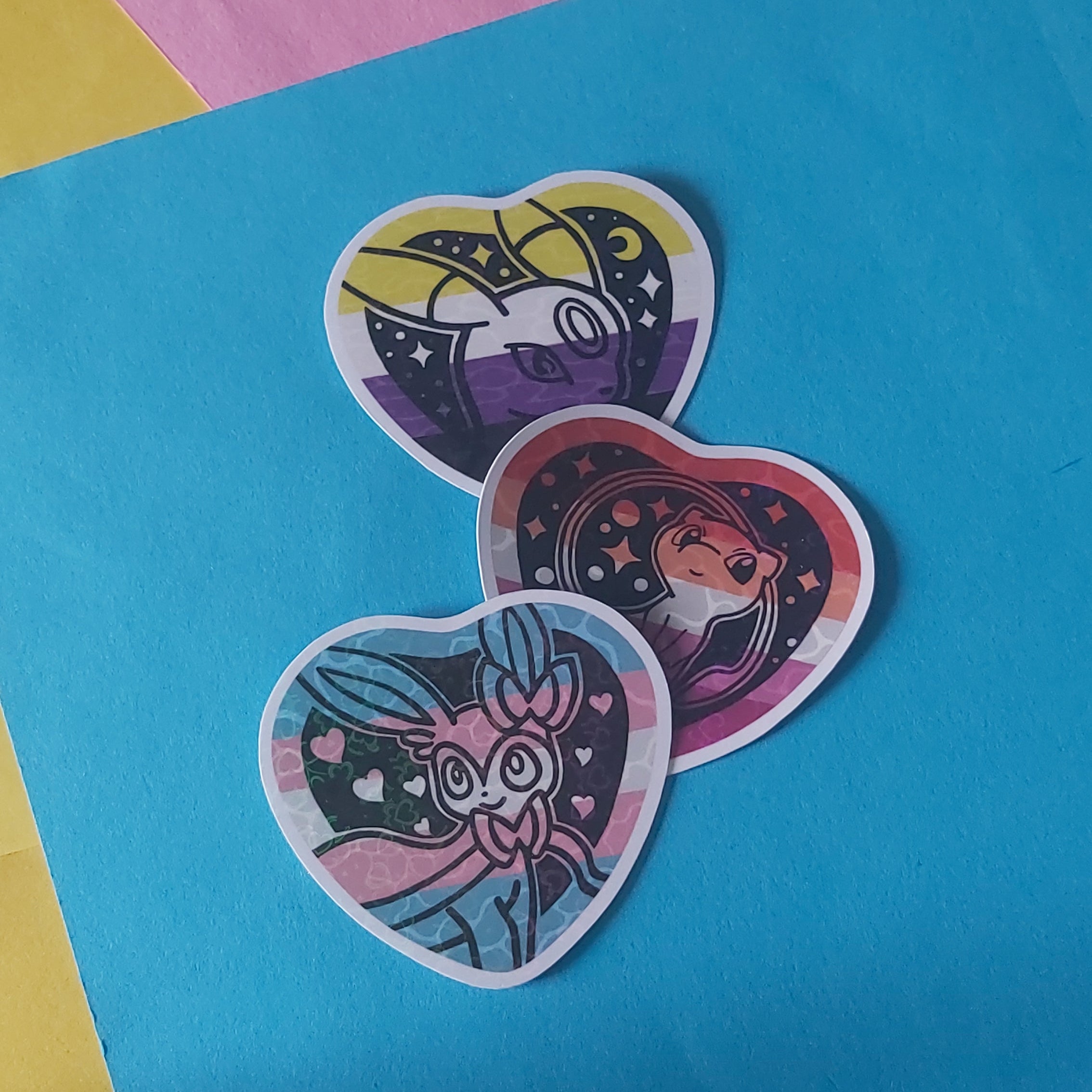 Three heart-shaped stickers featuring cartoon characters on a bright blue sheet of paper, with yellow and pink paper in the background.