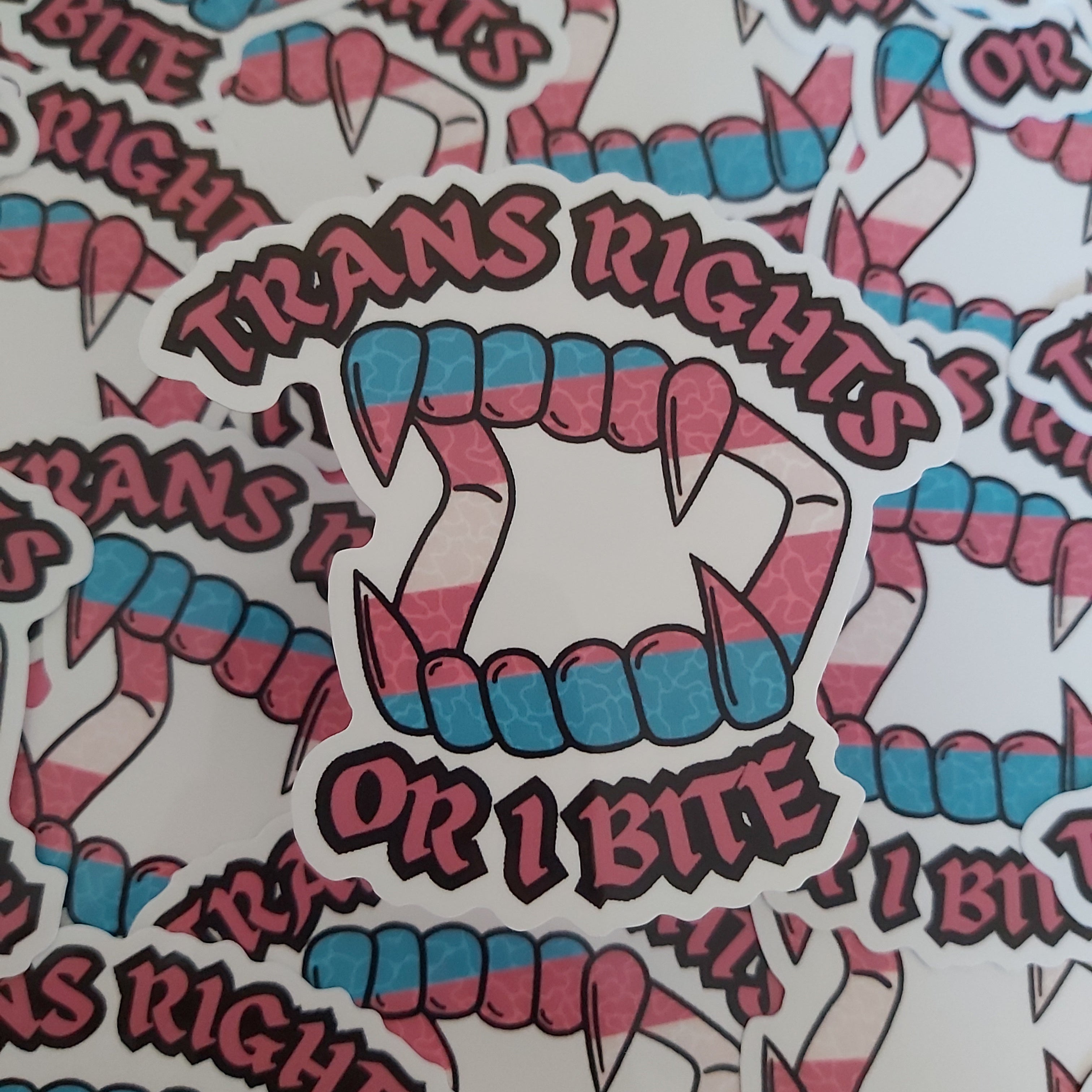 A close-up of a repeating sticker design featuring pink and blue teeth and fangs, plus the text 'TRANS RIGHTS OR I BITE' in stylised lettering.