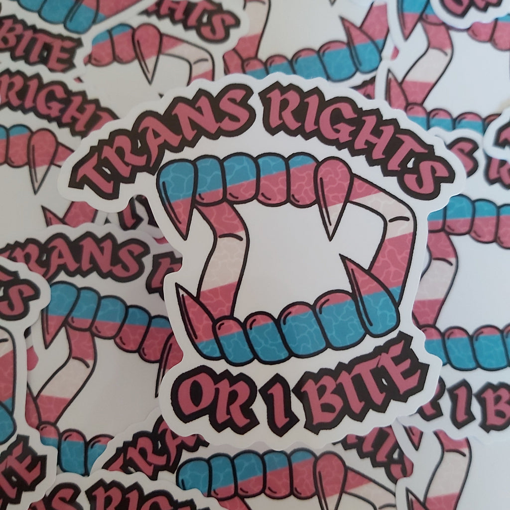 A close-up of multiple die-cut stickers featuring stylised fangs and the text 'TRANS RIGHTS OR I BITE' in pink and blue gradient lettering, arranged overlapping on a white background.