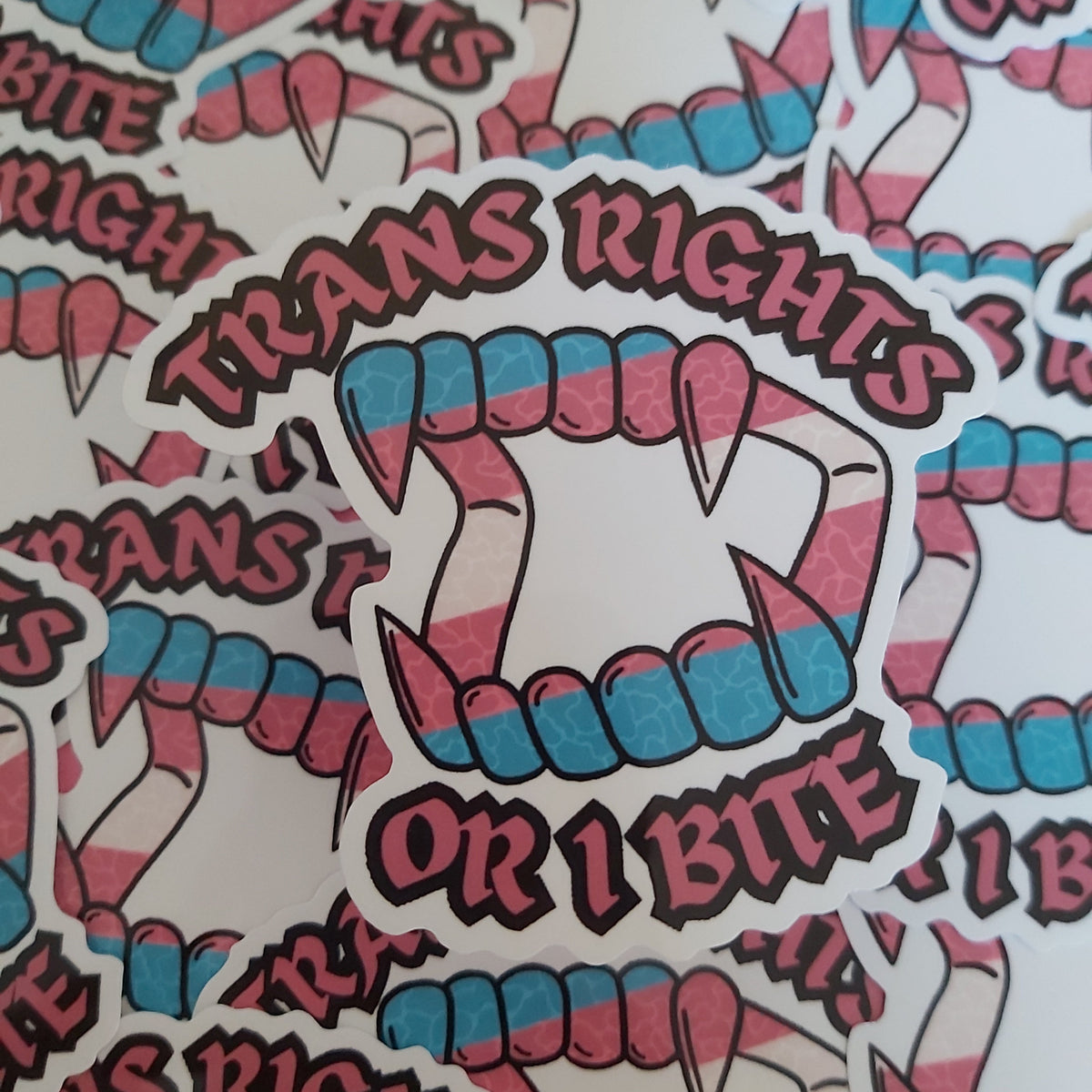 A close-up of multiple die-cut stickers featuring stylised fangs and the text 'TRANS RIGHTS OR I BITE' in pink and blue gradient lettering, arranged overlapping on a white background.