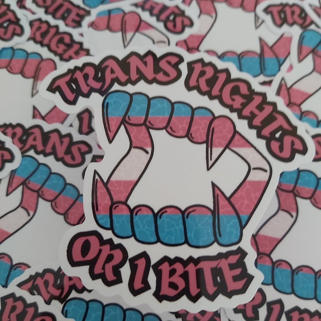 A close-up of multiple die-cut stickers featuring a vampire mouth with fangs and the text 'TRANS RIGHTS OR I BITE' in pink lettering, with the teeth colored in pink and blue stripes. The stickers are arranged on a white background.