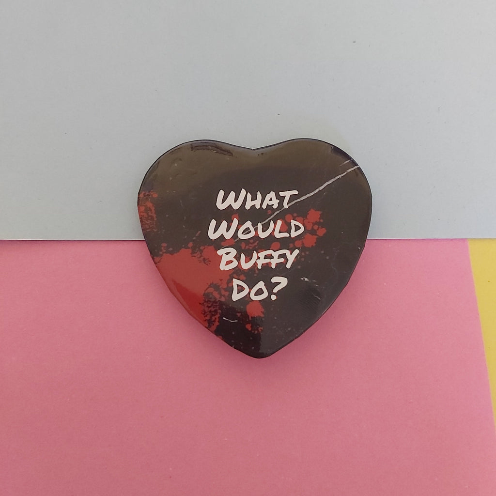 A heart-shaped black pin with white text reading 'WHAT WOULD BUFFY DO?' and red splatter details, displayed on a pink and blue background.