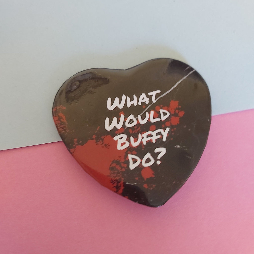 A heart-shaped pin with a black and red splatter design, featuring the white text 'WHAT WOULD BUFFY DO?' on a split blue and pink background.