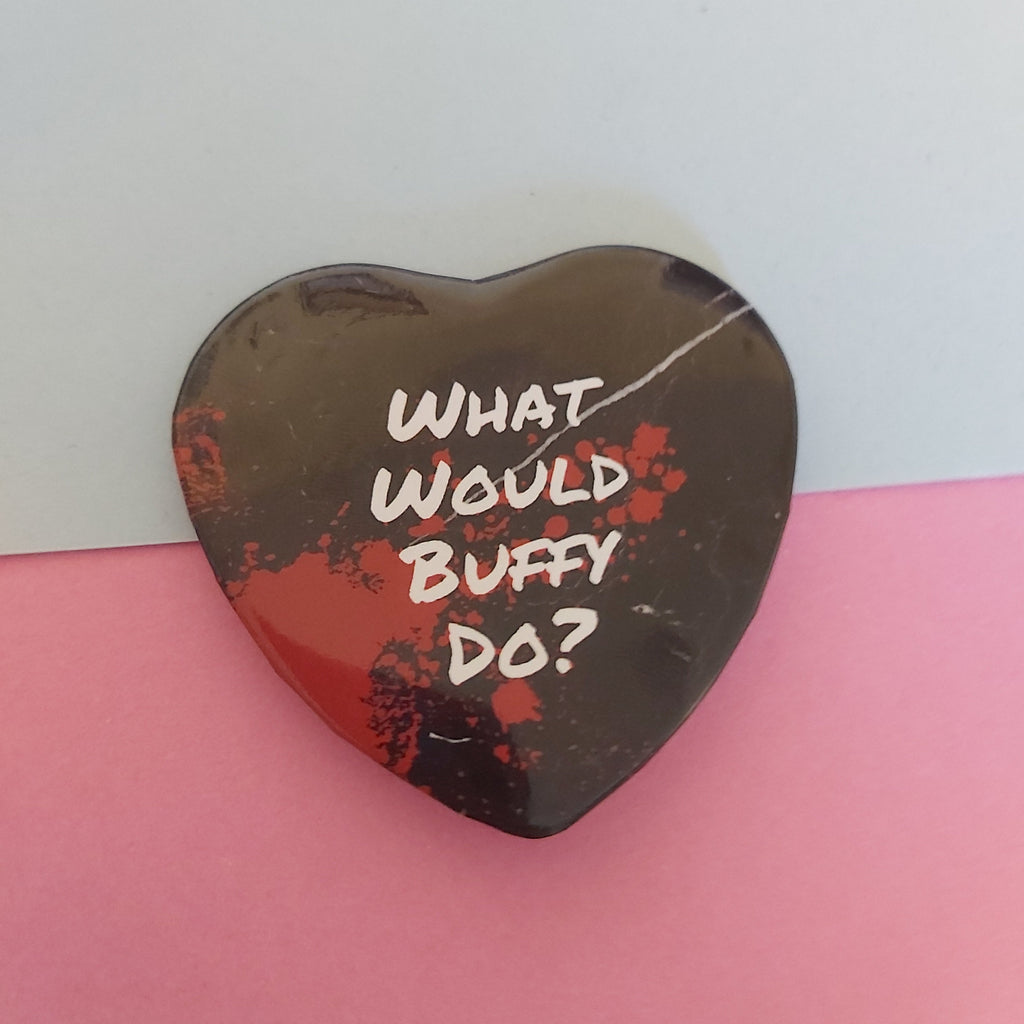 A heart-shaped pin with the text 'WHAT WOULD BUFFY DO?' in white, featuring a black marbled design with red splatter accents, set against a split blue and pink background.