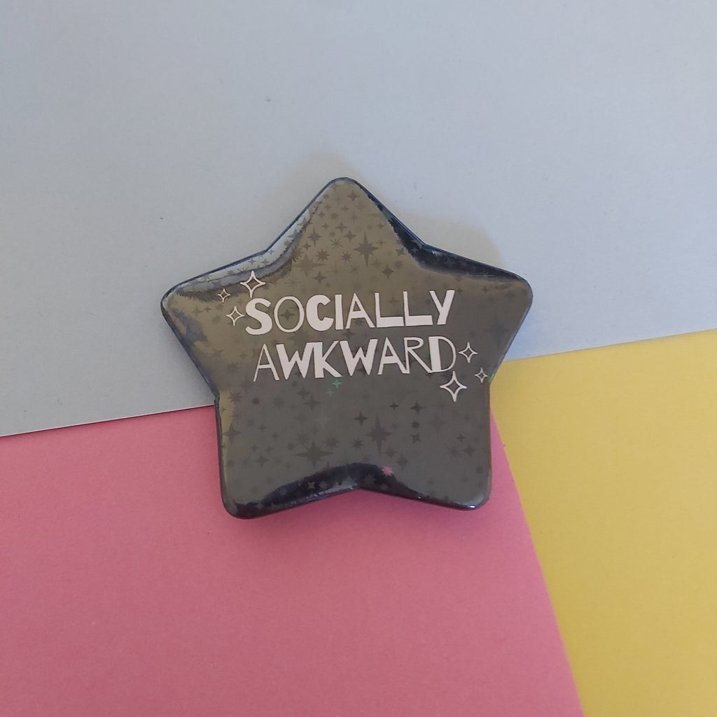 A star-shaped pin with the text 'SOCIALLY AWKWARD' and small sparkle accents, displayed on a background split diagonally into white, pink, and yellow sections.