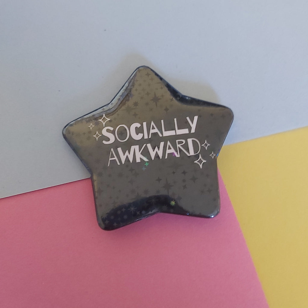 A star-shaped holographic pin with the words 'SOCIALLY AWKWARD' in white, placed on a background split diagonally into blue, pink, and yellow sections.