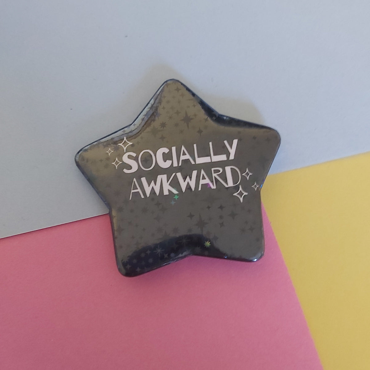 A star-shaped holographic pin with the words 'SOCIALLY AWKWARD' in white, placed on a background split diagonally into blue, pink, and yellow sections.