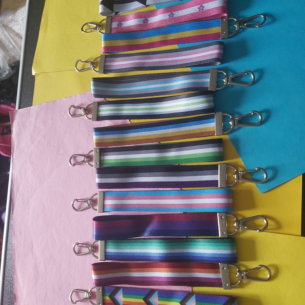 Ten colorful striped and patterned fabric lanyard straps with silver metal lobster clasps, arranged vertically on pink, yellow, and blue sheets of paper.