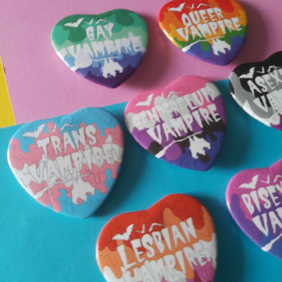 A set of heart-shaped pins featuring various LGBTQ+ themed vampire designs, displayed on a split pink and blue background.