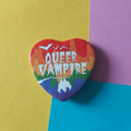 A heart-shaped pin featuring a rainbow paint background and the text 'QUEER VAMPIRE' in white, displayed on a diagonally split yellow, pink, and blue background.