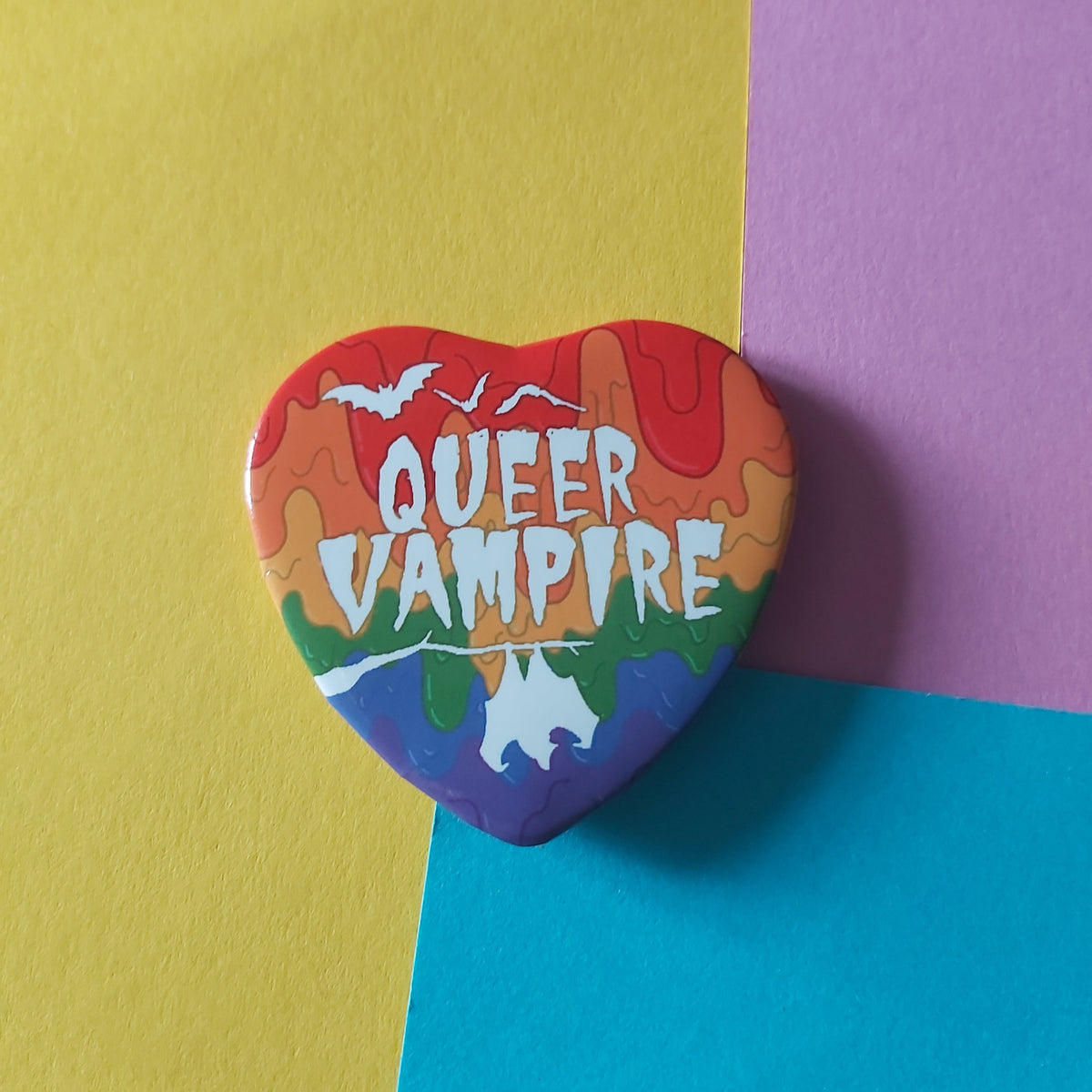 A heart-shaped pin featuring a rainbow paint background and the text 'QUEER VAMPIRE' in white, displayed on a diagonally split yellow, pink, and blue background.