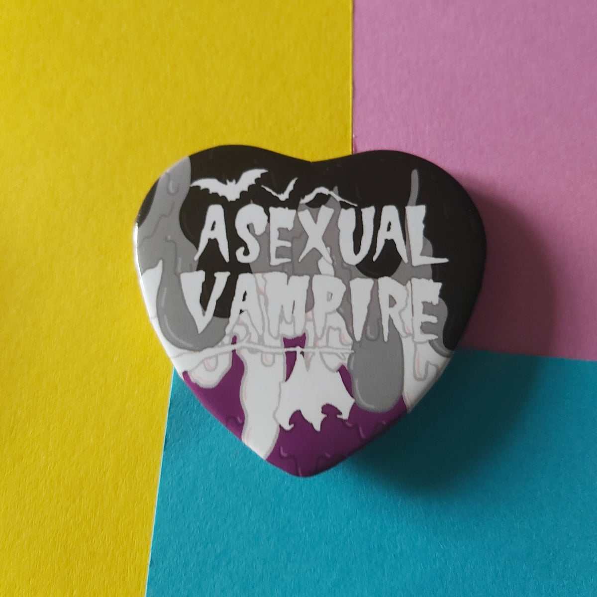 A heart-shaped pin with a black, gray, white, and purple gradient design featuring the text 'ASEXUAL VAMPIRE' and bat silhouettes, displayed on a background of yellow, pink, and blue paper.