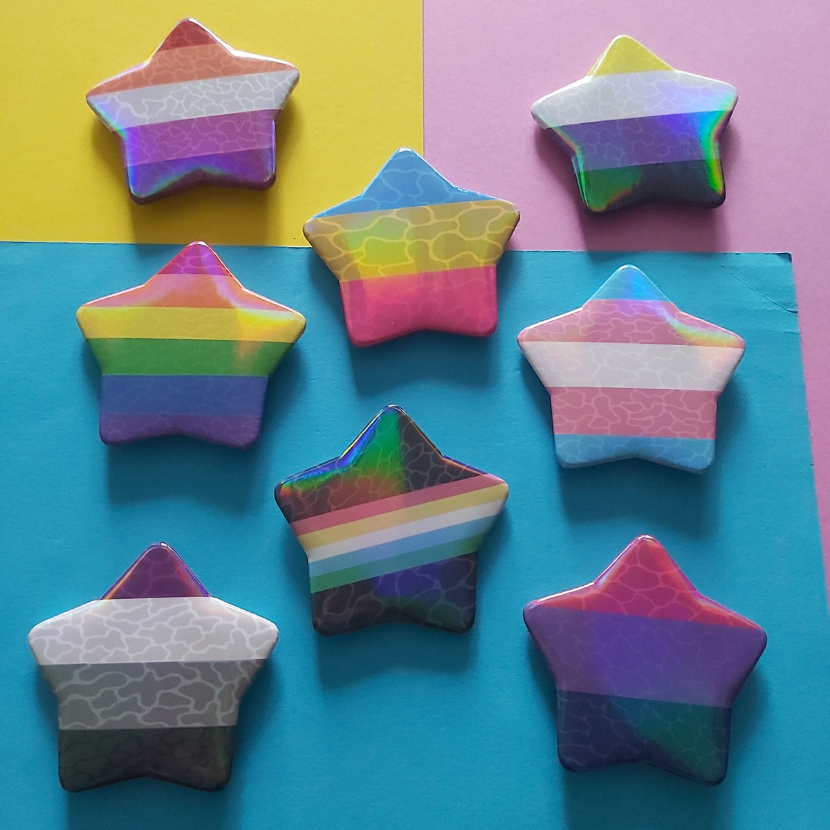 Eight star-shaped pins with various pride flag patterns, arranged on a colourful background of yellow, pink, and blue paper.