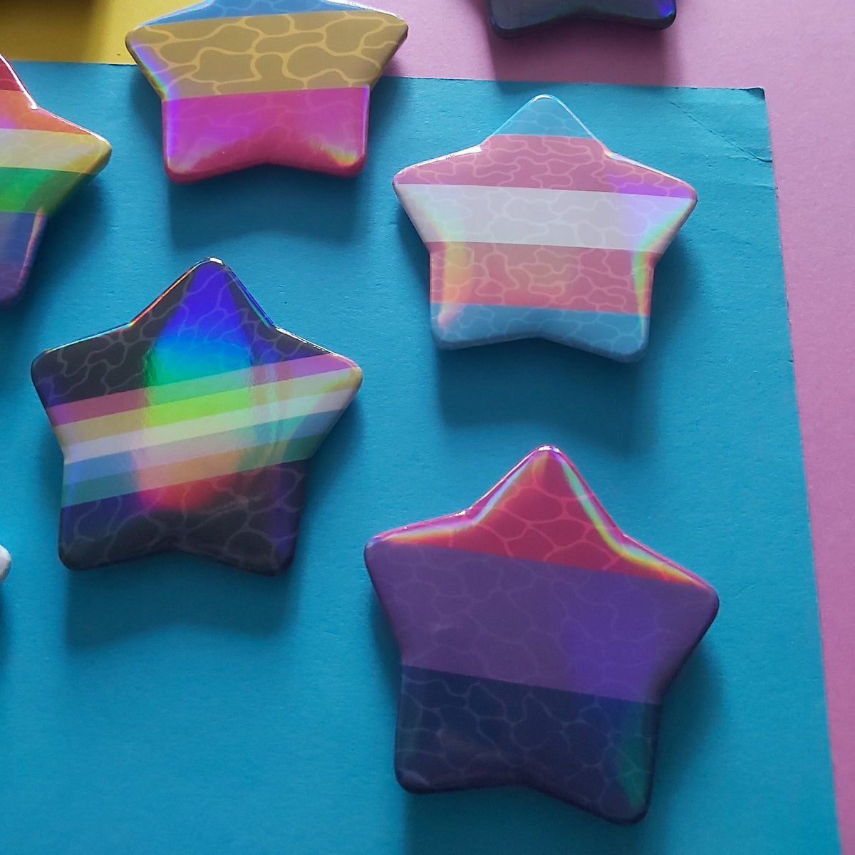 A collection of star-shaped pins with various pride flag designs and textured finishes, arranged on a background of yellow, pink, and blue paper.