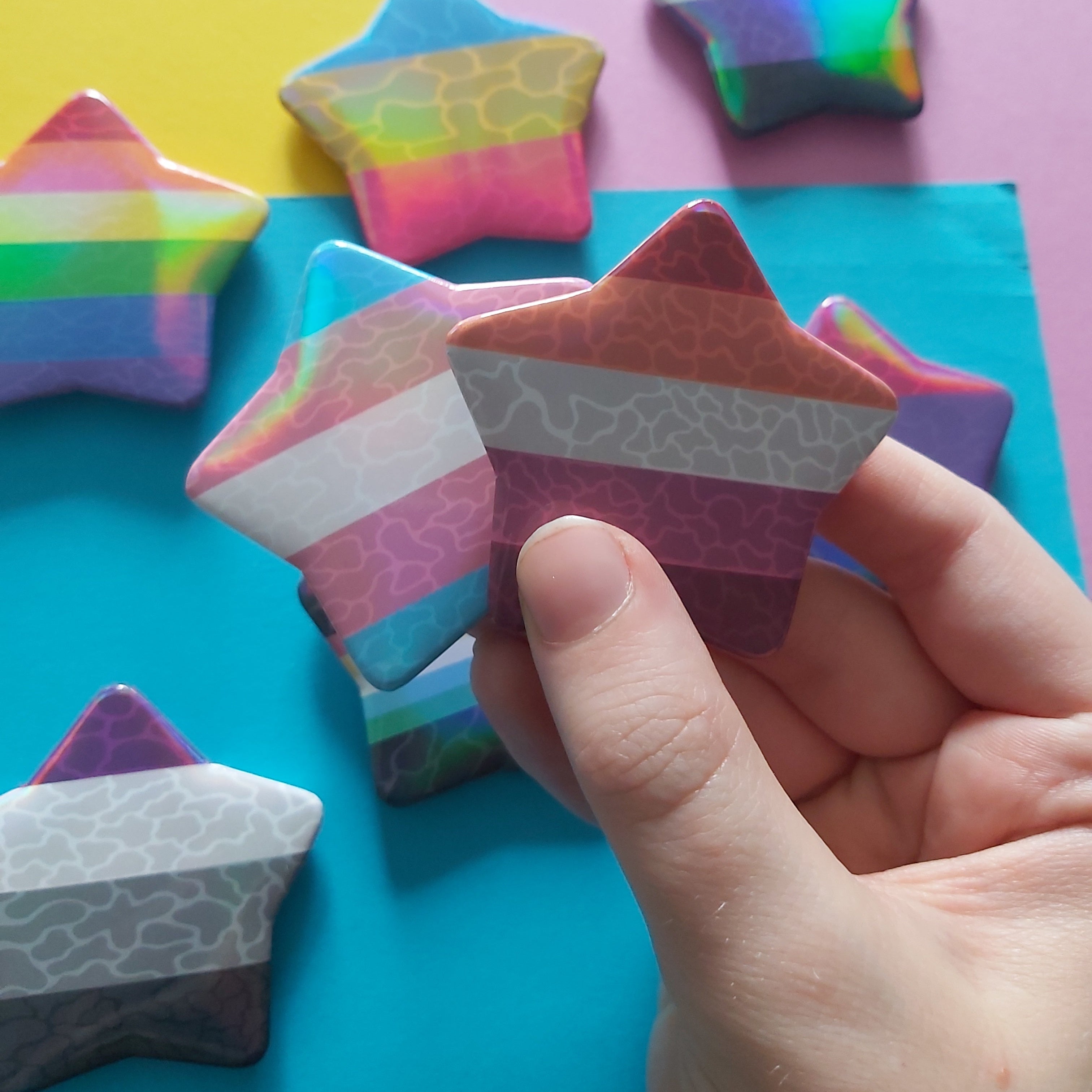 A hand holds two star-shaped pins, one with the transgender pride flag and the other with the lesbian flag, with other star pins in various colours scattered in the background on a split yellow and blue surface.