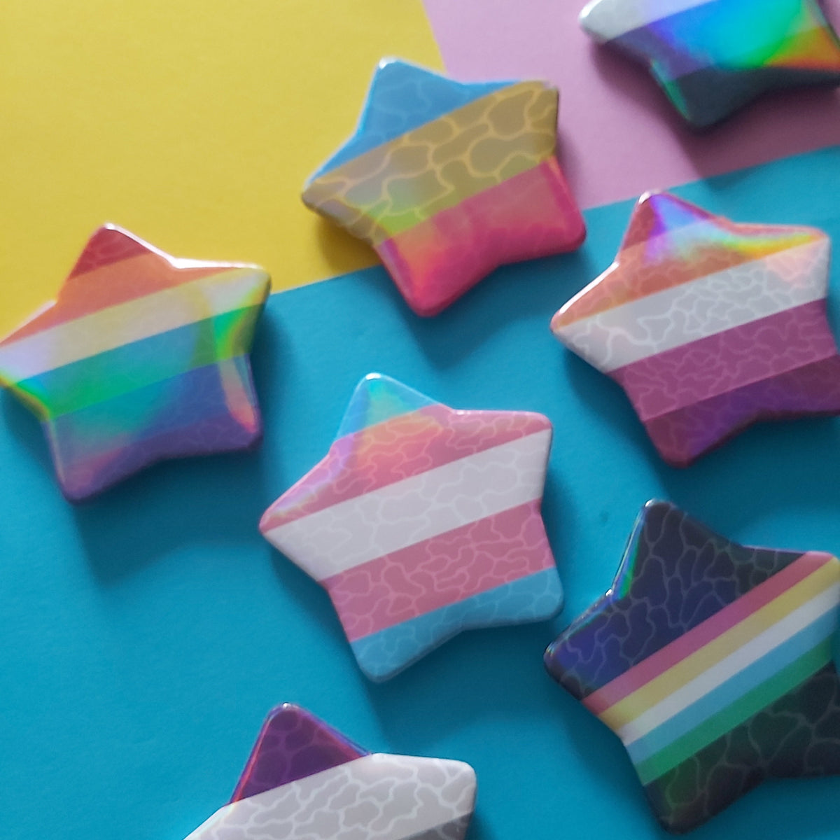 A set of star-shaped pins with various pride flag designs, including rainbow, bisexual, pansexual, lesbian, transgender, and non-binary stripes, arranged on a colorful background of yellow, pink, and blue.