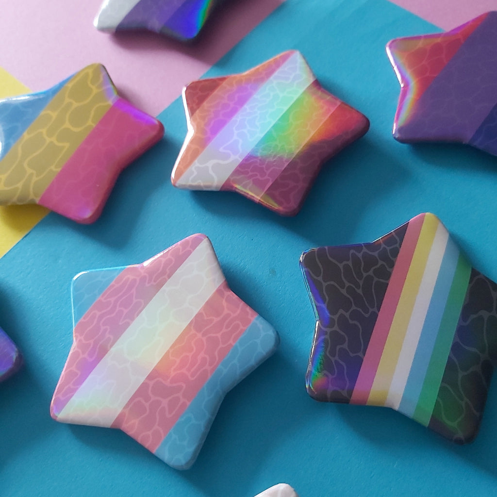 A collection of star-shaped pins with various pride flags, arranged on a split pink and blue background.