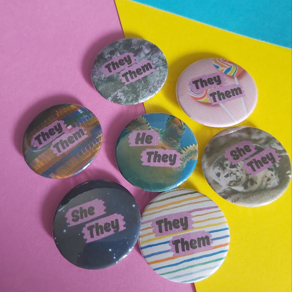 Seven round pinback buttons with pronoun designs are arranged on a diagonally split pink and yellow background with a blue corner.