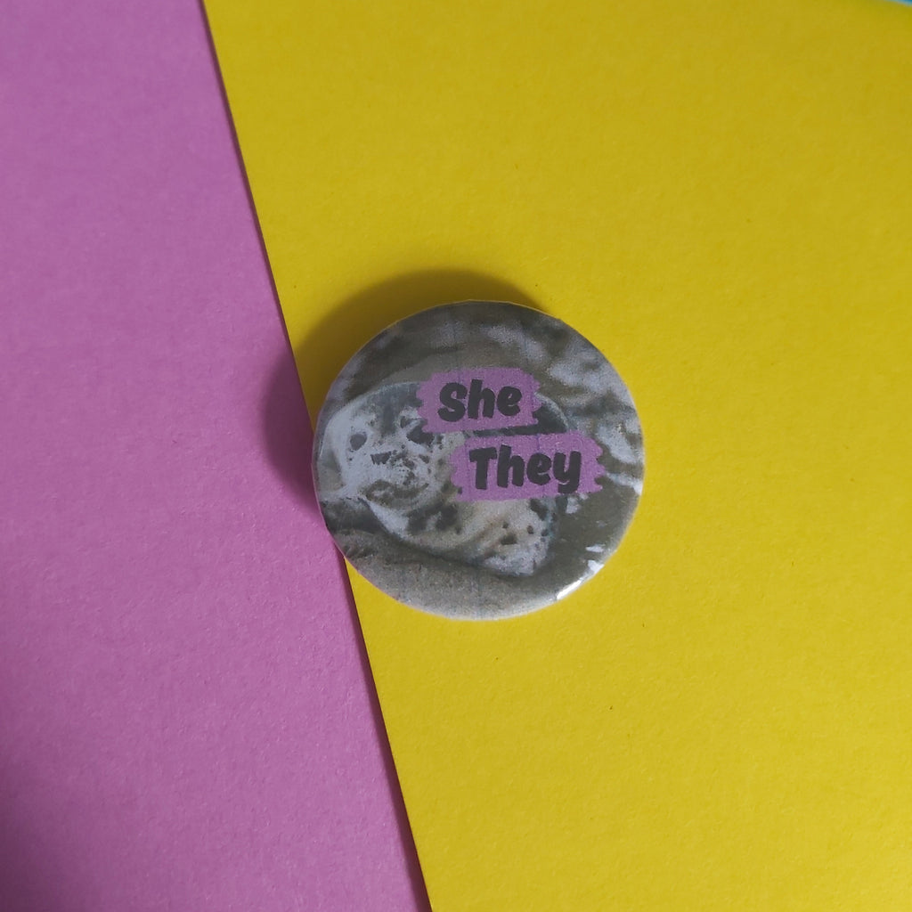 A round pinback button badge featuring a black and white seal photo that reads 'She - They', placed on a background of pink, yellow, and blue paper.