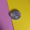 A round pinback button badge featuring a black and white seal photo that reads 'She - They', placed on a background of pink, yellow, and blue paper.