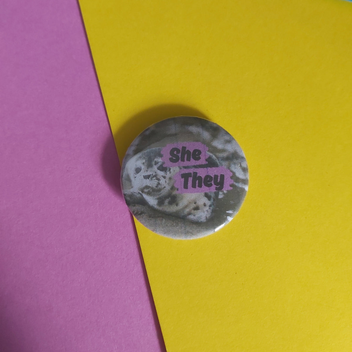 A round pinback button badge featuring a black and white seal photo that reads 'She - They', placed on a background of pink, yellow, and blue paper.