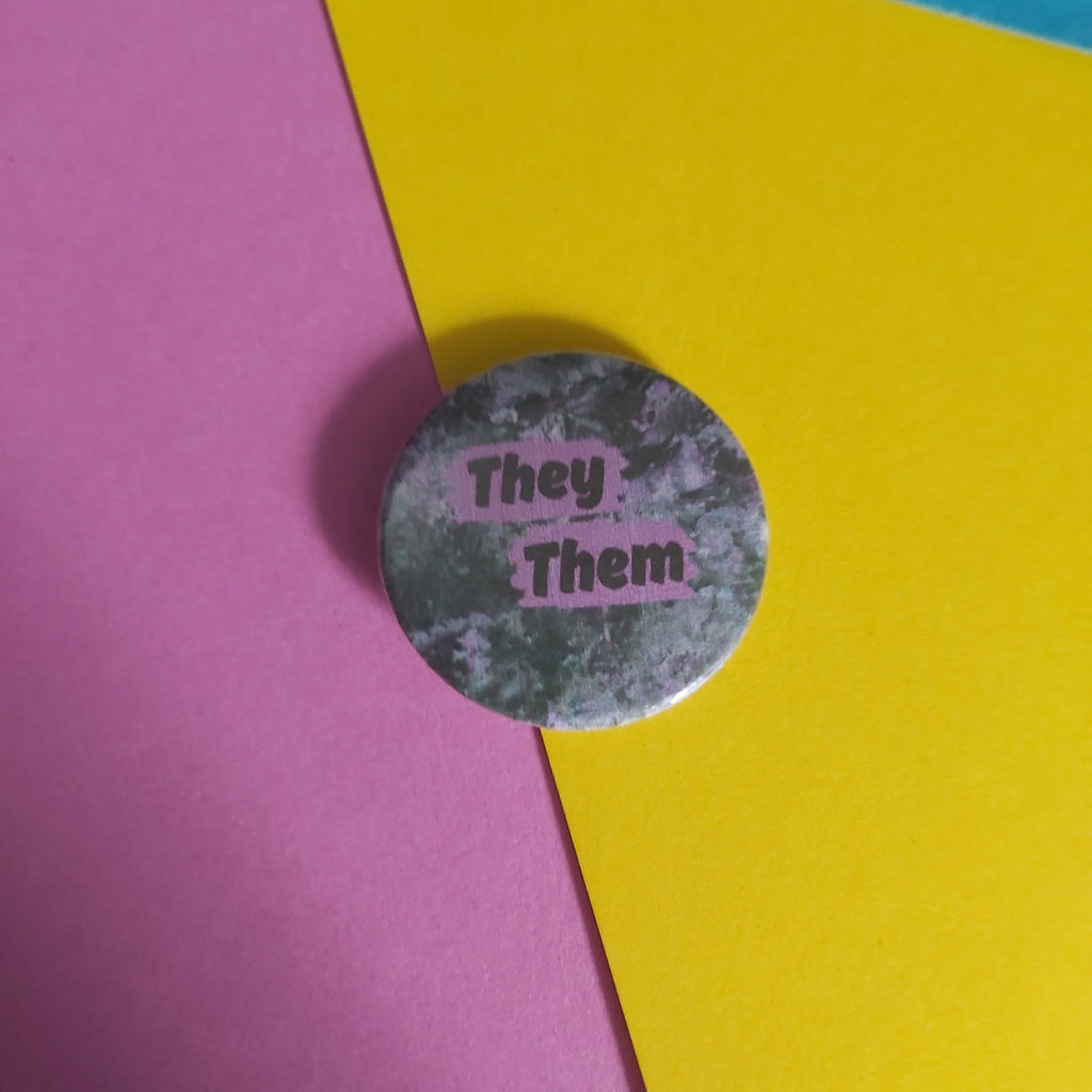A round pinback button badge with a distressed gray and green camouflage pattern and pink text reading 'They Them', placed on a background of diagonally split yellow, pink, and blue paper.