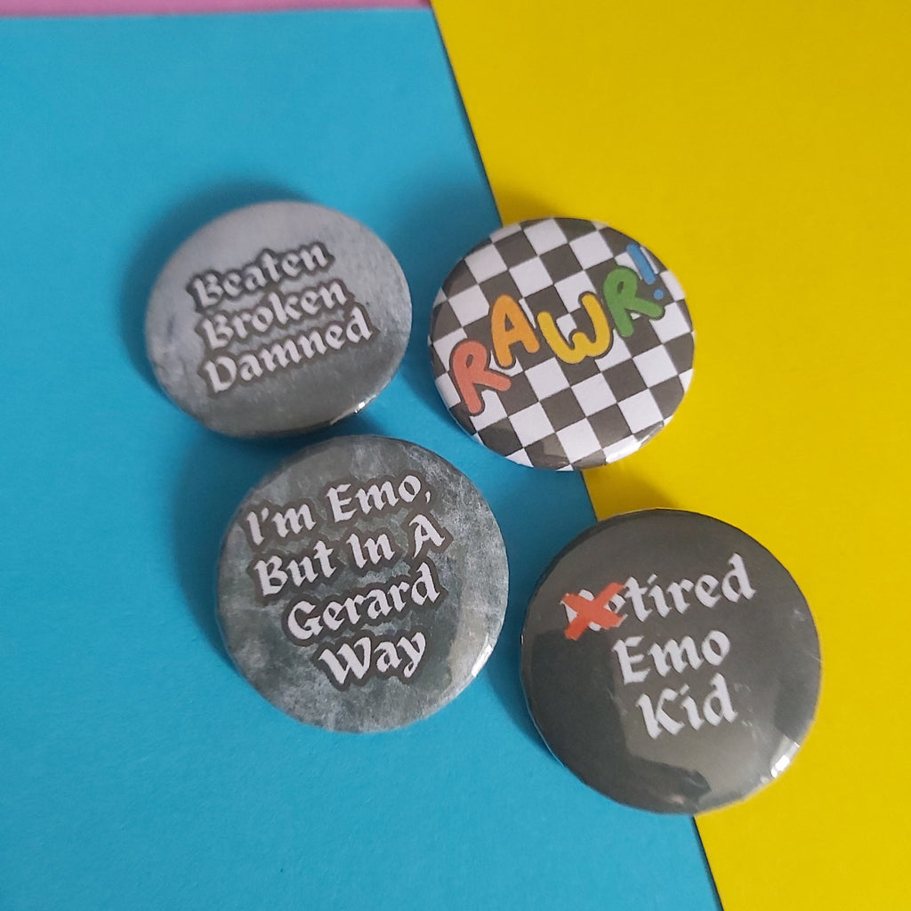 Four round pin-back buttons with humorous text designs arranged on a colourful background of pink, blue, and yellow paper. The buttons are grey with white text; one features a black-and-white checkerboard pattern with the word 'RAWR' in multicolour letters, and one says 'Retired Emo Kid'
