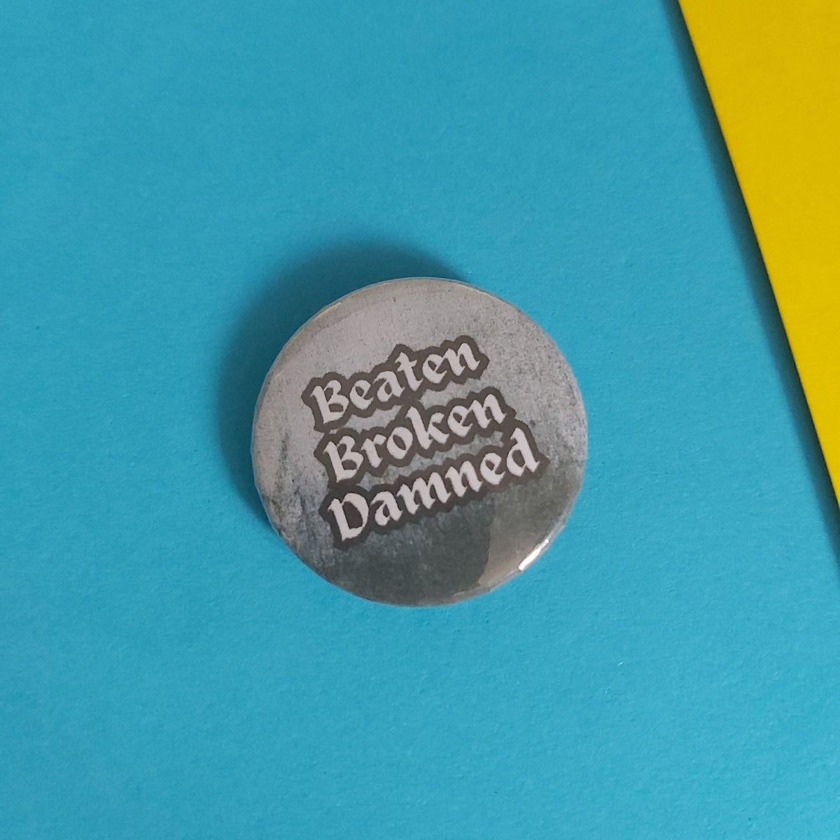 A round grey pinback button with the words 'Beaten Broken Damned' in white gothic-style lettering, placed on a bright blue background with a yellow shape in the top right corner.
