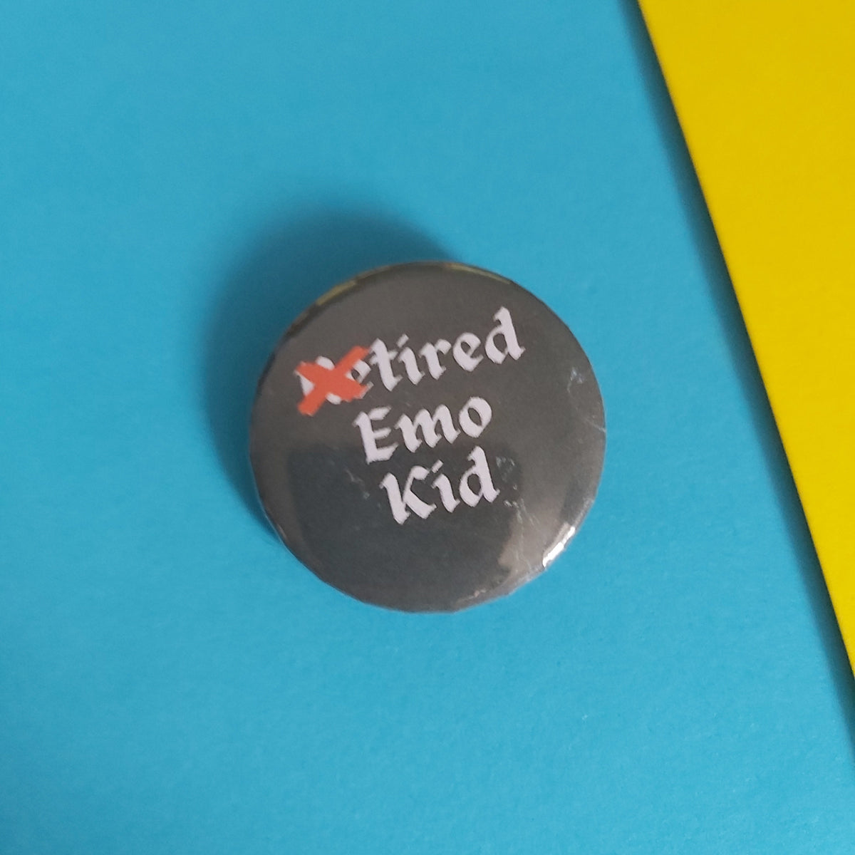 A round pinback button with the text 'Retired Emo Kid' in white, featuring a red 'X' through 'RE', shown on a bright blue background with a yellow corner visible.