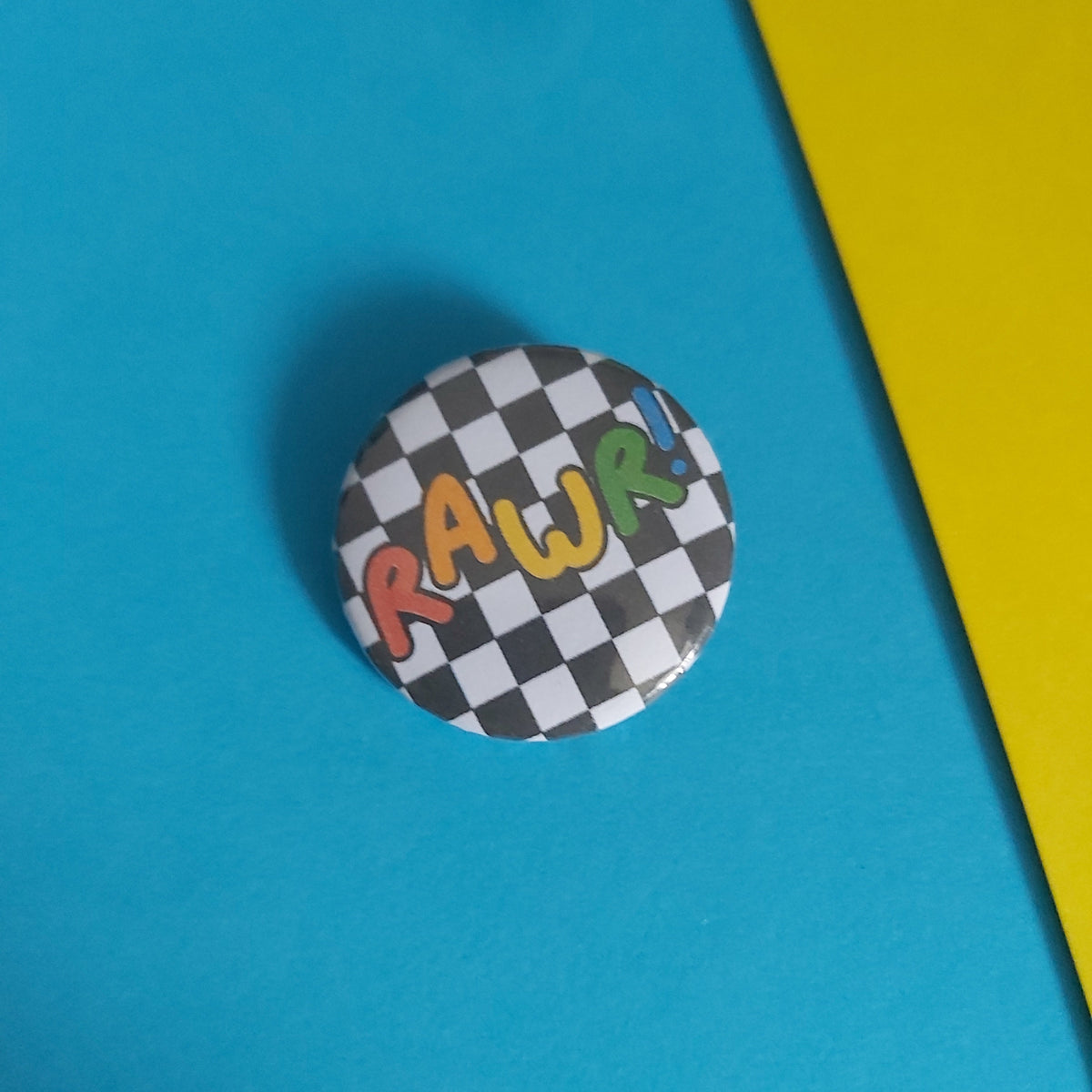 A round pinback button badge featuring a black and white checkered pattern background with the word 'RAWR' in colorful, playful letters, resting on a bright blue surface with a yellow corner visible.