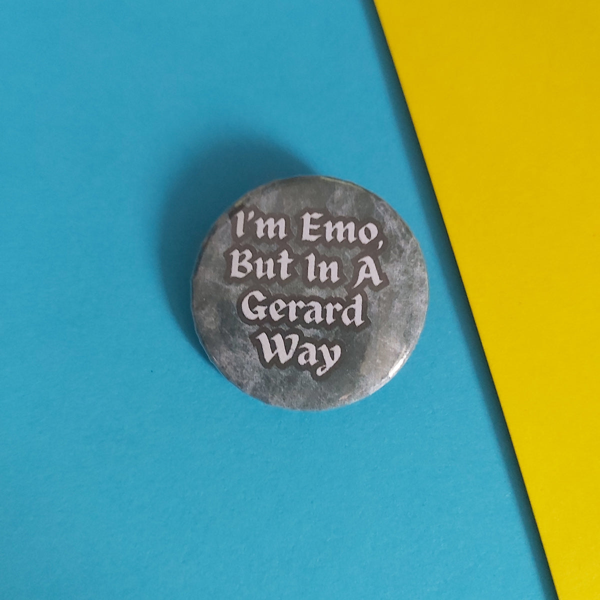 A round gray pinback button with the text 'I'm Emo, But In A Gerard Way' in white, placed on a diagonally split blue and yellow background.