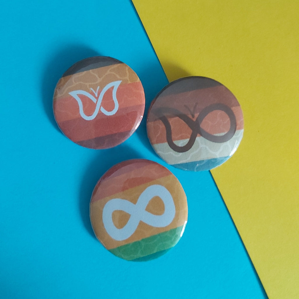 Three small round pin-back buttons with Neurodivergent Pride flag stripes and white symbols (a butterfly and an infinity symbol) arranged on a diagonally split blue and yellow background.