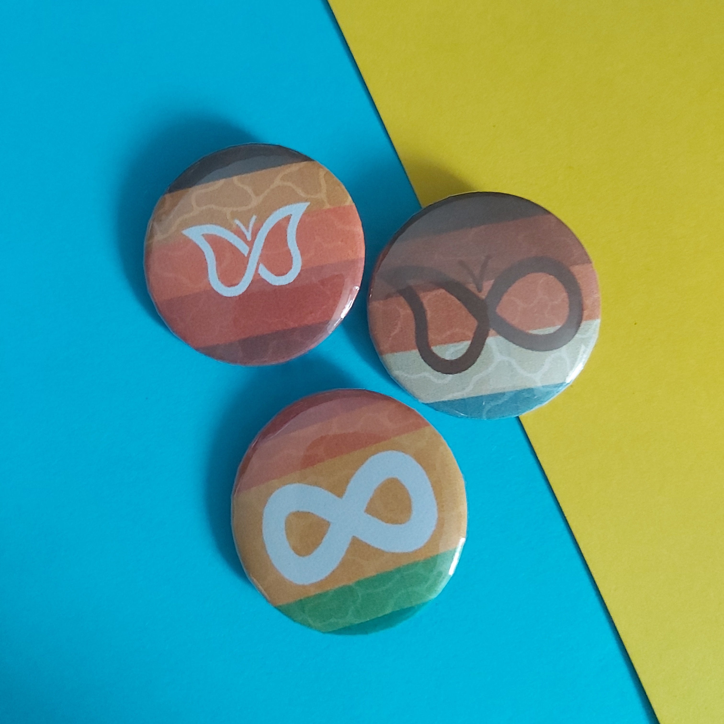 Three small round pin-back buttons with a striped background and white symbols (a butterfly, a loop, and an infinity symbol), arranged on a diagonally split bright blue and yellow surface.