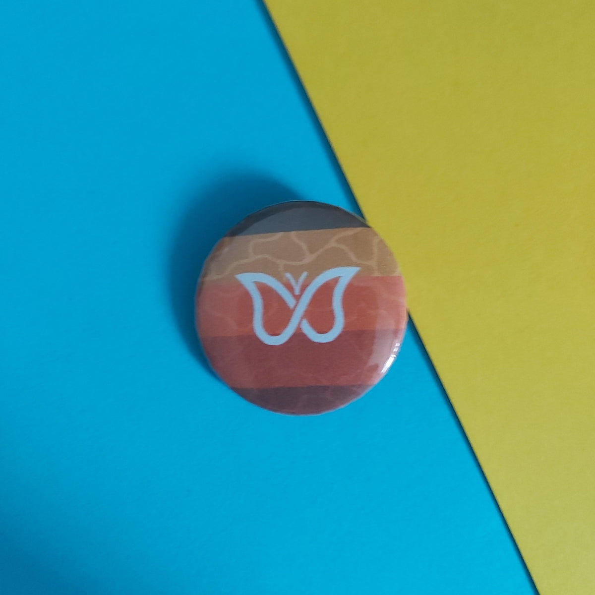 A round pinback button featuring a white butterfly logo over horizontal stripes in brown, orange, and red, placed on a diagonally split blue and yellow background.